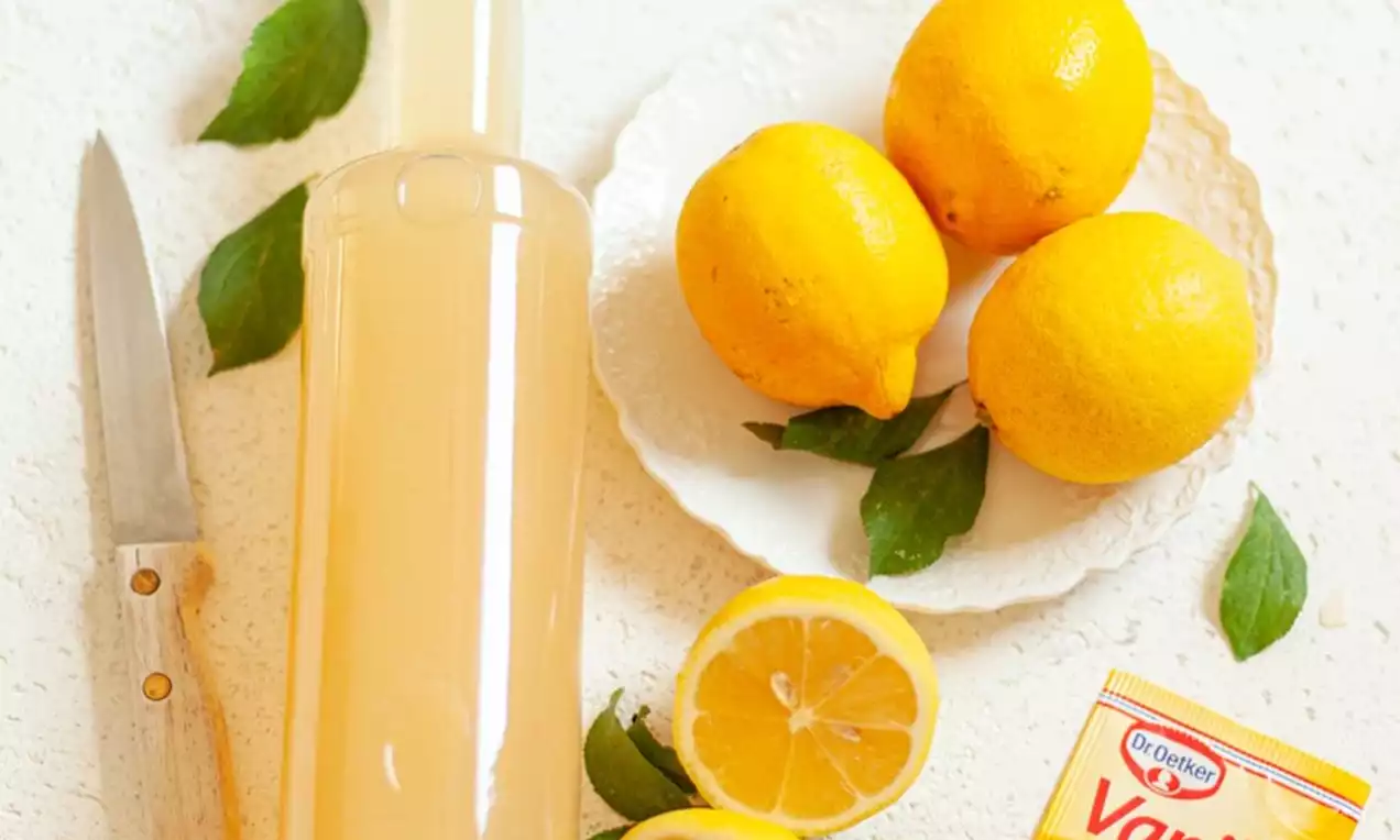 Prozirna staklena boca ispunjena blijedožutim likerom, okružena limunima, listovima, nožem i paketićem vanilina na svijetloj podlozi