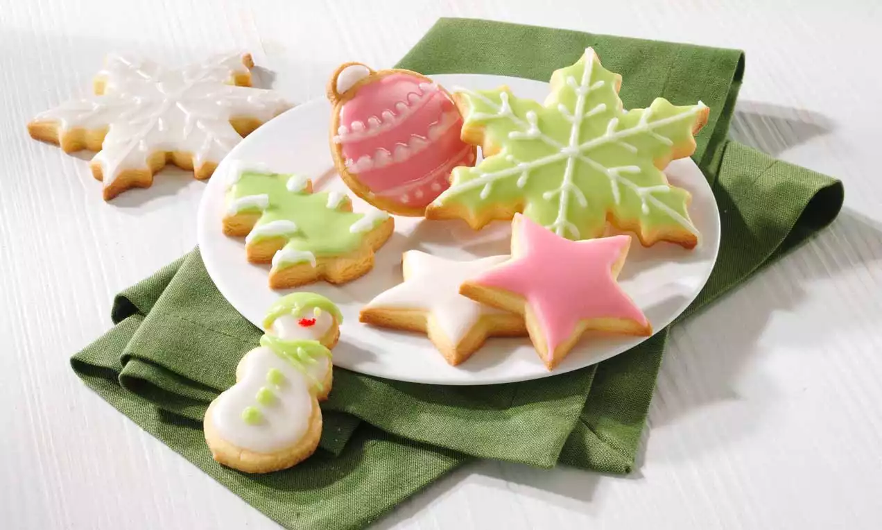 Auf einem weißen Teller liegen bunte Weihnachts-Butterkekse in Stern-, Schneeflocken-, Baum- und Schneemannform auf grüner Stoffserviette