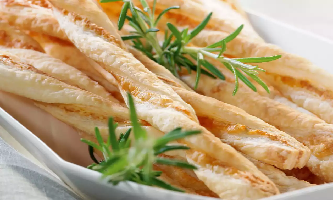 Croccanti bastoncini dorati di pasta sfoglia intrecciata con lievi venature, disposti su un piatto bianco guarniti con rametti di rosmarino зелен