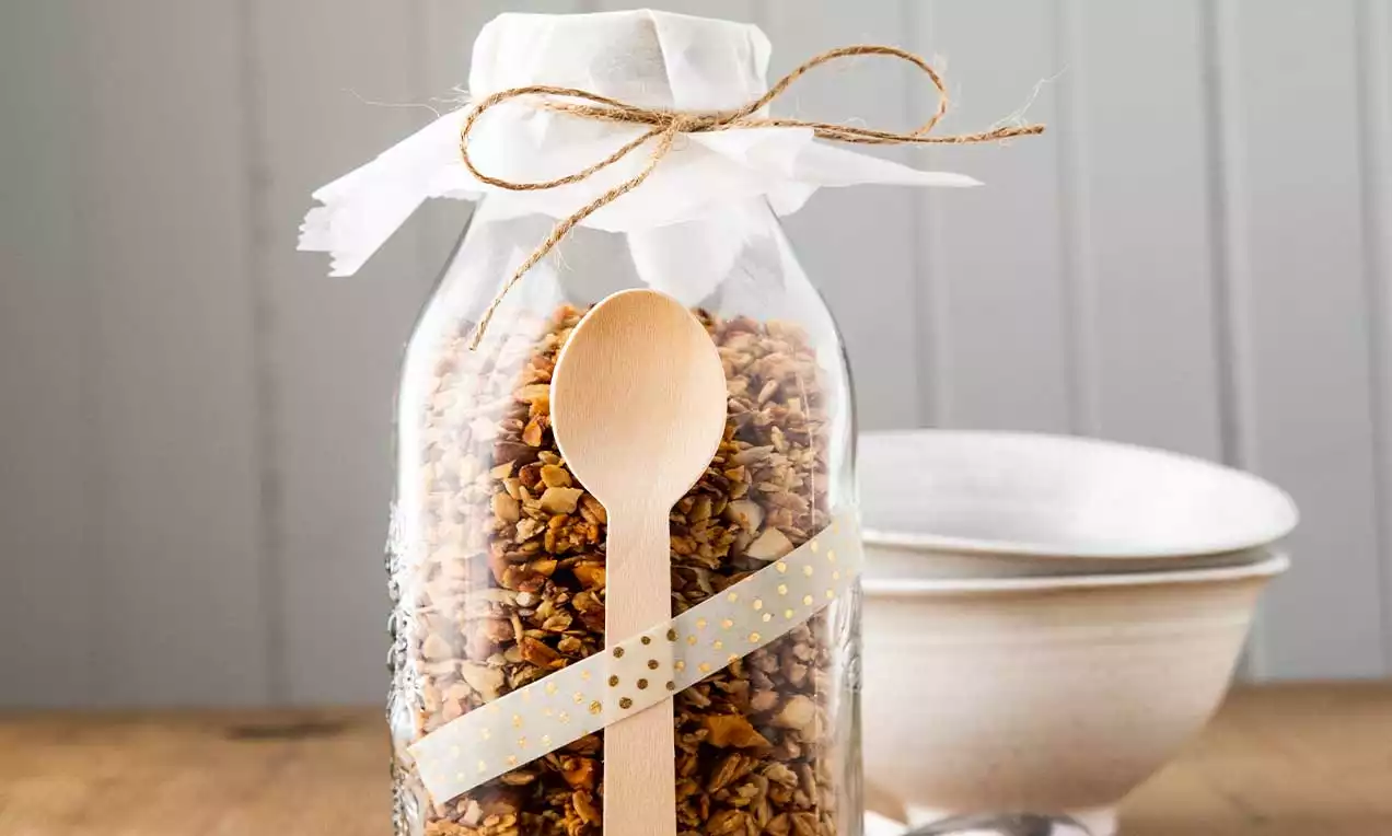Granola dorata dai toni nocciola in un barattolo di vetro con cucchiaio di legno decorato, avvolto da un nastro beige su un tavolo di legno