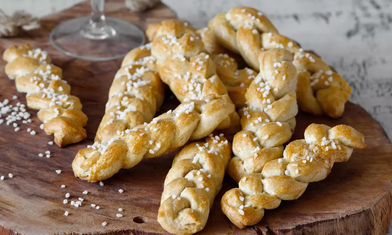 Zlatavé pečené copánky s jemně lesklým povrchem, posypané krystalky cukru, uspořádané volně na rustikálním dřevěném podnosu