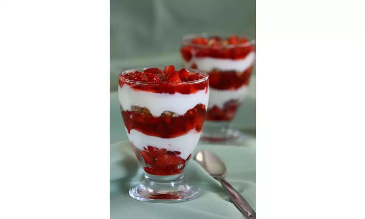 Cam kadehte kat kat yerleştirilmiş canlı kırmızı çilek parçaları ve beyaz yoğurt, yanında metal bir kaşık ile açık yeşil fon önünde