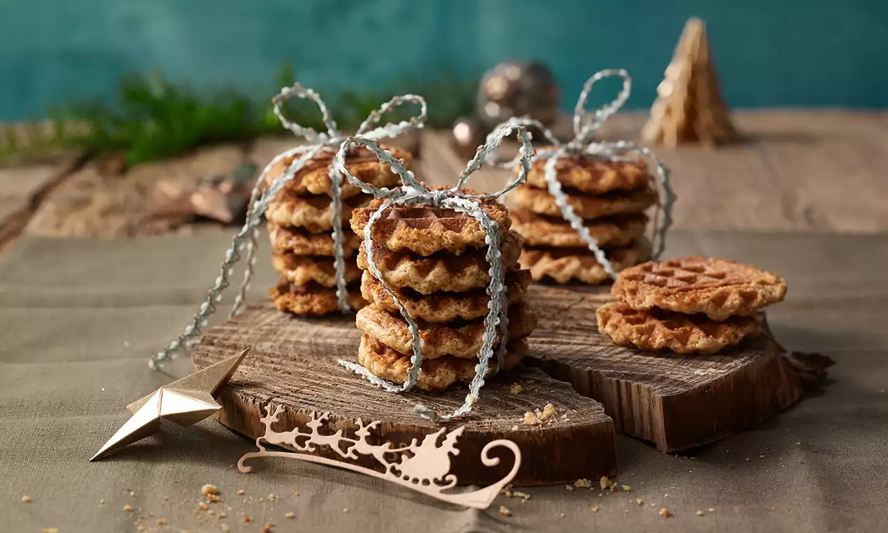 Knusprige, braune Waffelkekse mit eingebackener Gewürzstruktur, auf rustikalem Holzbrett gestapelt und silbernen Schleifen verziert