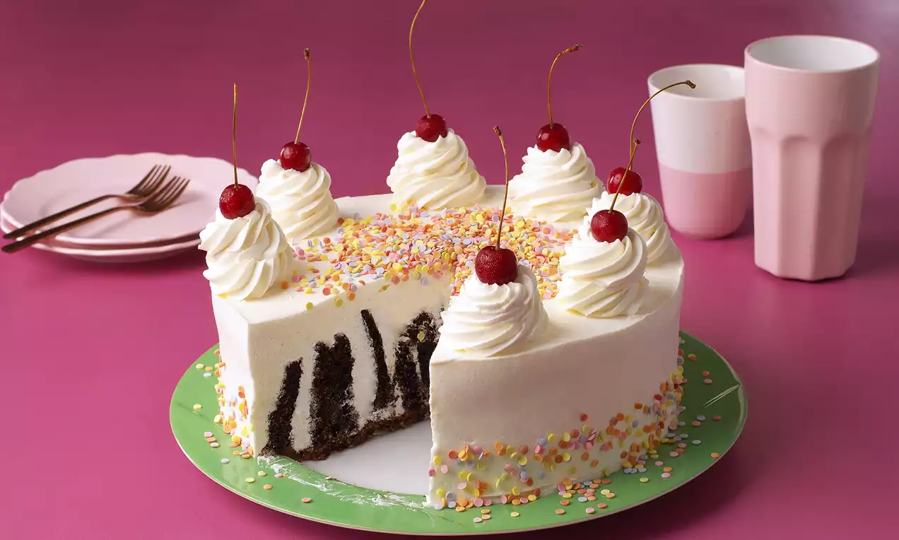 Beyaz kremalı yuvarlak pasta, içi dikey kakaolu katmanlı, üstü krem şanti ve kirazlarla süslenmiş, renkli şekerdekorlarla çevrili