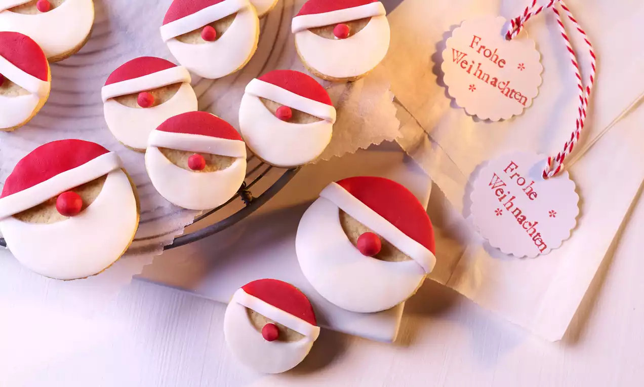 Keksi okruglog oblika ukrašeni bijelim fondantom, crvenom kapicom od marcipana i kuglicama, na pergamentu i žičanoj rešetki