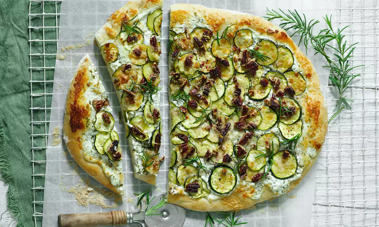 Cuketová pizza so zlatistým okrajom, zelenými plátkami cukety, hnedými kúskami orechov, bylinkami a krémovou bielou základňou na bielom pergamene