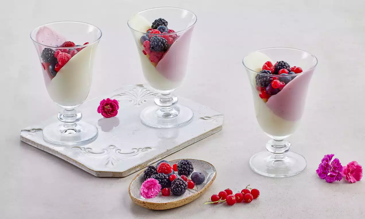 Şeffaf cam kupalarda eğimli beyaz ve pembe krema, üstünde böğürtlen, frambuaz ve kırmızı kuş üzümü, yanlarında çiçek detayları