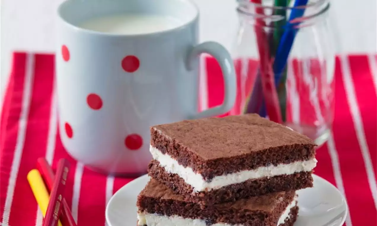 Dva obdélníkové řezy s tmavě hnědou vláčnou kakaovou vrstvou a krémovou bílou náplní na bílém talíři na červené kostkované utěrce