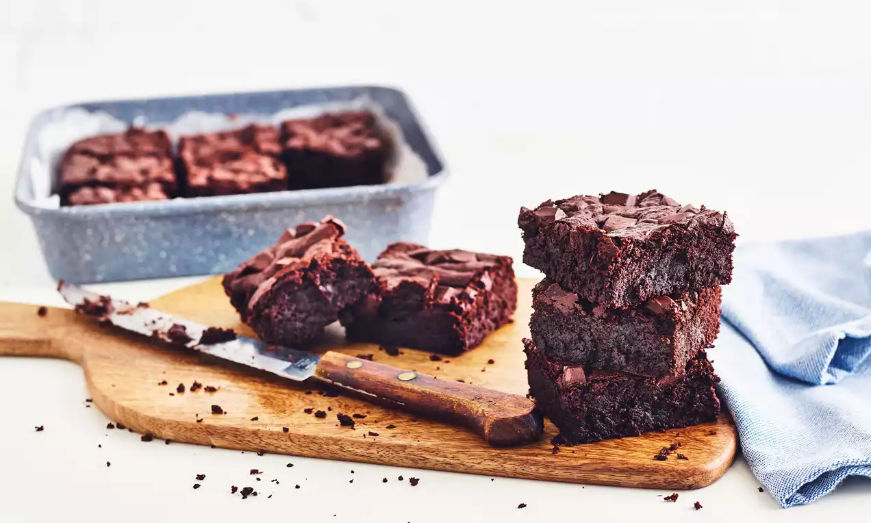 Rich, dark brown vegan brownies with a dense, fudgy texture stacked and scattered on a wooden board next to a tray and knife