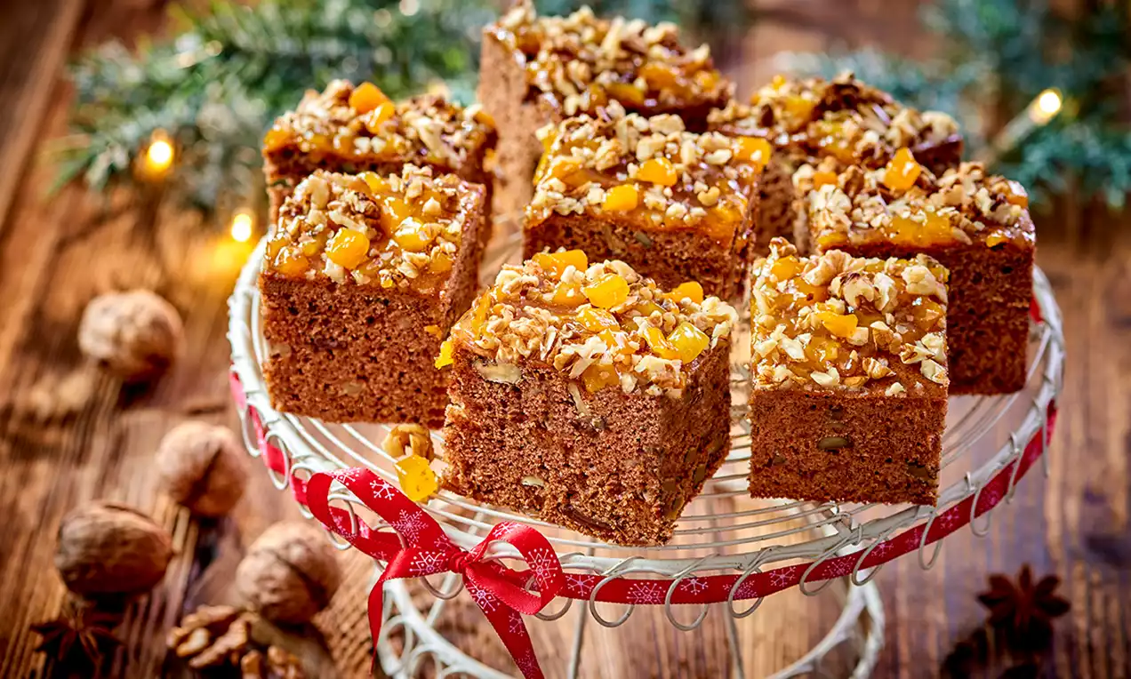 Kwadraty złocistobrązowego piernika z bakaliami i kawałkami moreli na ozdobnym talerzu z czerwoną wstążką, w tle gałązki jodły i orzechy