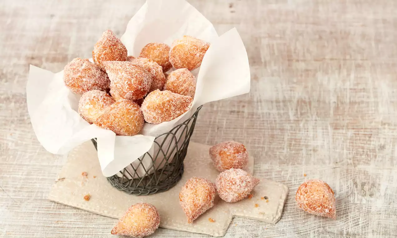 Goldbraune, zuckerbestäubte Muzenmandeln mit knuspriger Textur in einem Drahtkorb mit weißem Papier, einige auf hellen Holzunterlagen verstreut