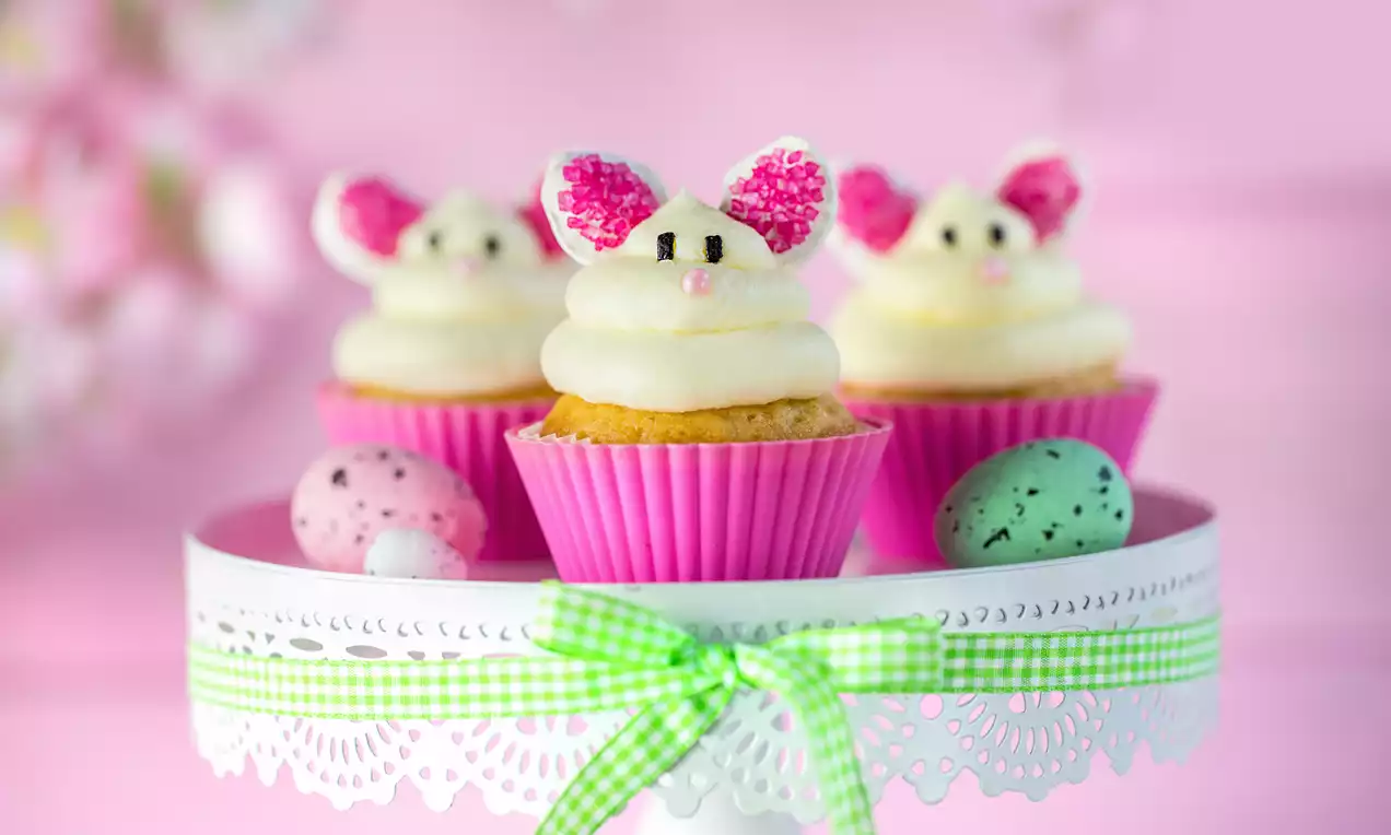 Babeczki waniliowe w różowych papilotkach z kremowym białym lukrem, dekorowane cukrowymi uszami i oczkami, na białej paterze z zieloną wstążką