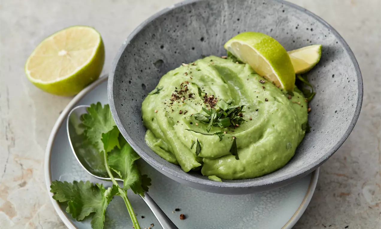 Cremige, grün gefärbte Guacamole in einer grauen Schale mit frischen Kräutern, Limettenspalten und schwarzem Pfeffer auf einem Teller angerichtet