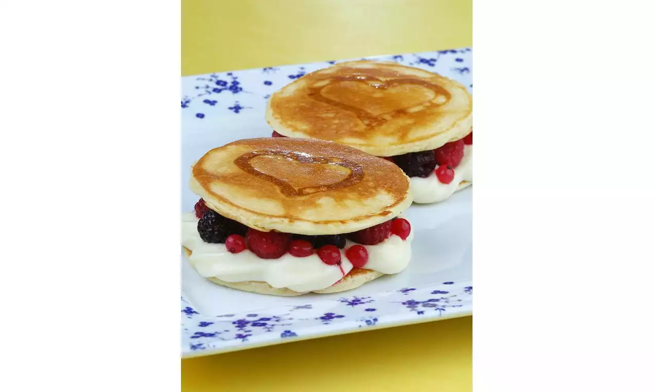 Kalp şekilli desene sahip yumuşak altın rengi pancakeler arasında beyaz krema, kırmızı ve siyah meyvelerle porselen çiçekli tabakta