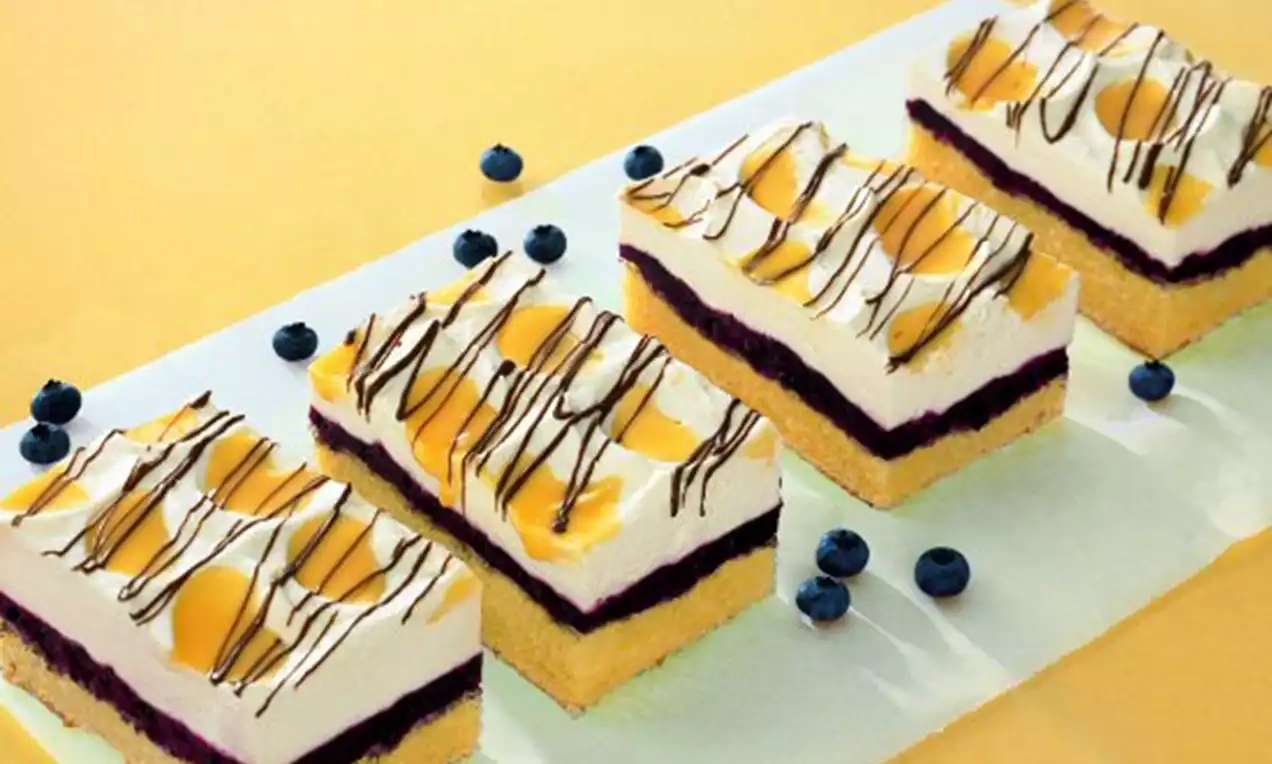 Rechteckige Kuchenstücke mit hellem Boden, dunkler Heidelbeerfüllung, heller Creme, Pfirsichhälften und Schokostreifen, dekoriert mit Heidelbeeren