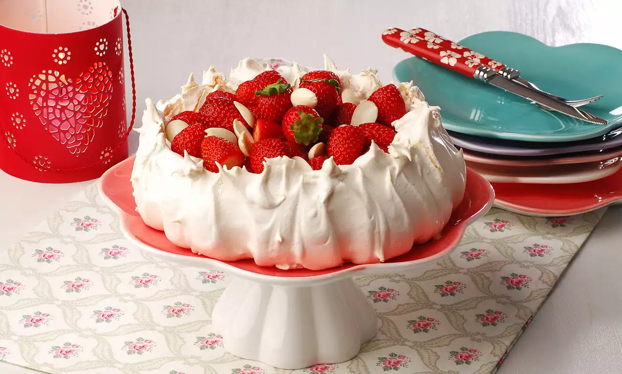 Krem beyaz beze tabanlı, ortası taze çilek ve badem dilimleriyle dolu, kırmızı tabakta romantik bir sunumla servis edilmiş tatlı