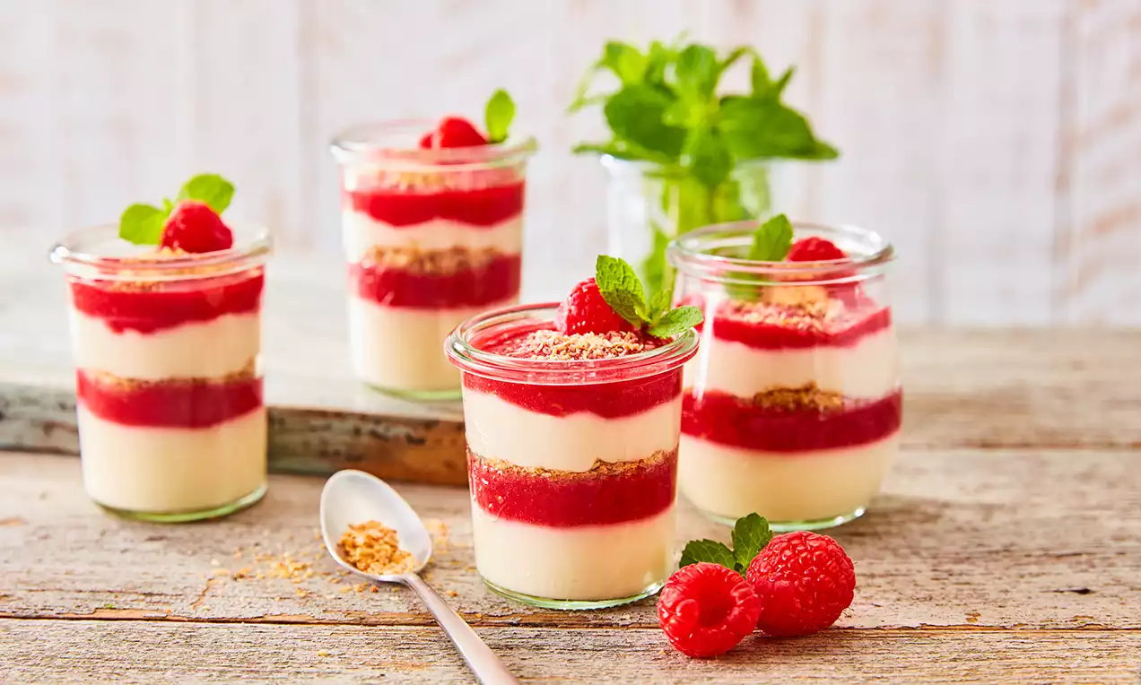 Mehrschichtiger veganer Kokospudding in Gläsern, abwechselnd mit rotem Himbeerpüree, dekoriert mit frischen Himbeeren und Minzblättern