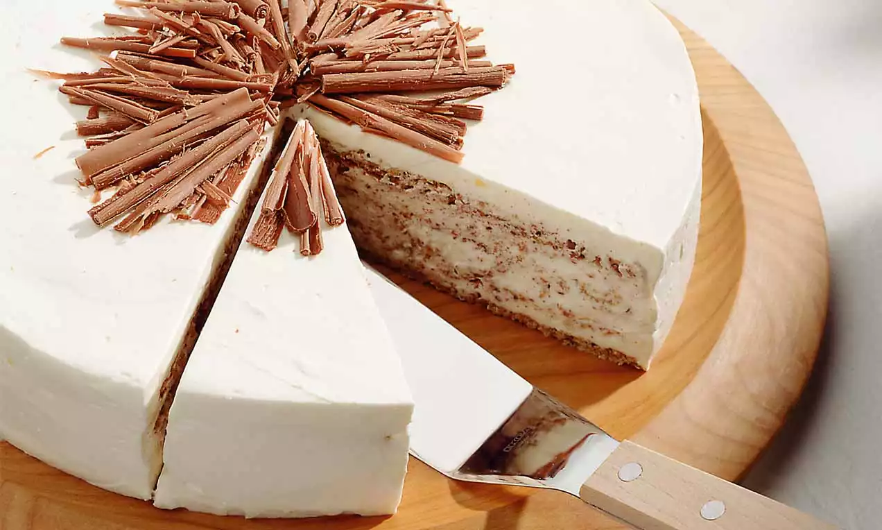 Schwedentorte mit weißer Glasur, feinen Schokoschichten, dekoriert mit Schokospänen, auf einem runden Holzbrett mit Kuchenheber