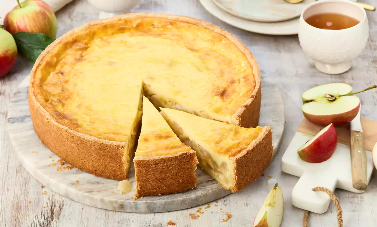 Apfel-Schmand-Kuchen mit goldgelber Füllung und knusprigem Rand auf einer Marmorplatte, dekoriert mit frischen Äpfeln und gedecktem Tisch