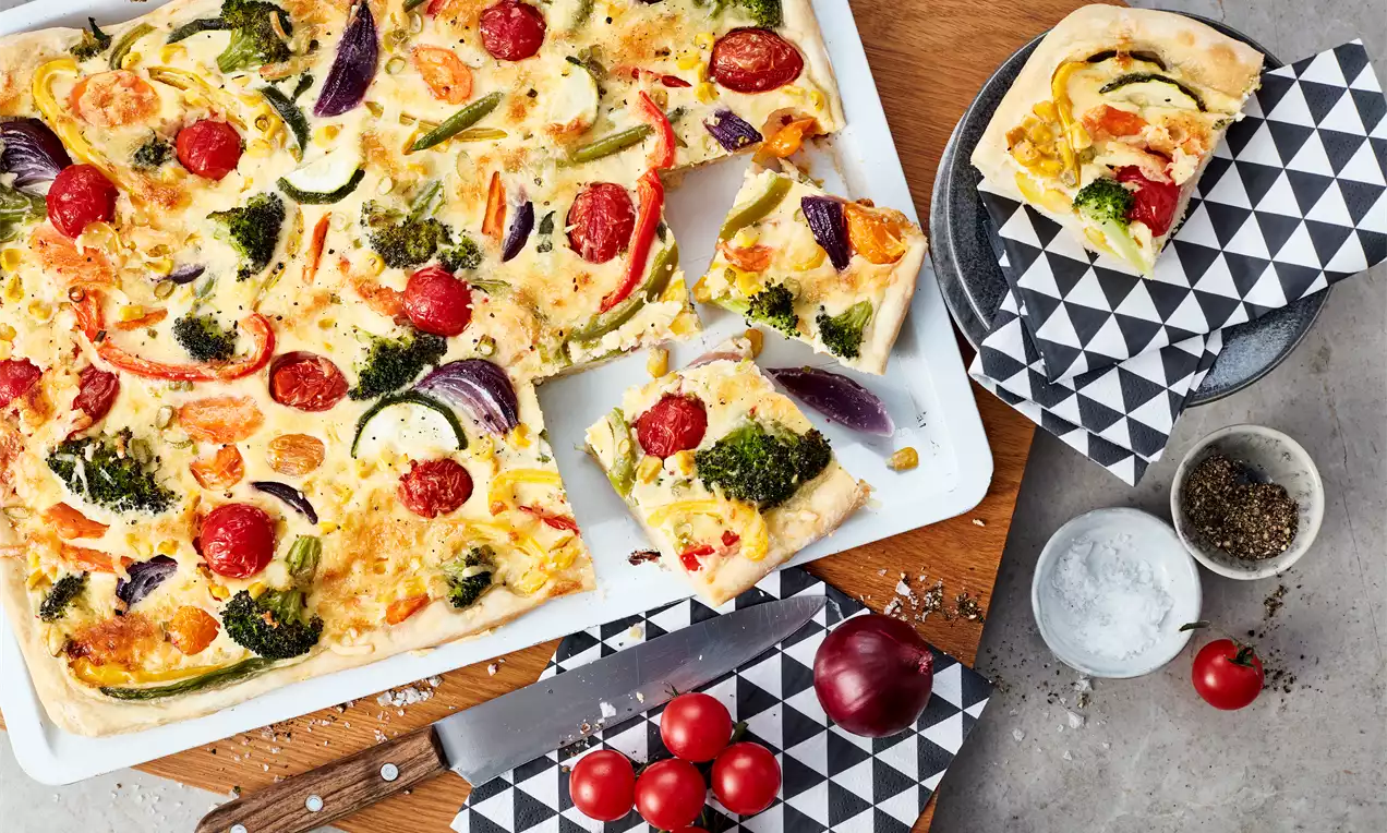Bunter Gemüsekuchen mit roten Tomaten, grünen Brokkolistückchen und hellgelbem Käse auf weißem Blech, mit einem Stück auf geometrierter Serviette