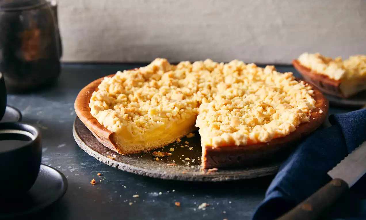 Crumble taart met lichtgele banketbakkersroom en kruimelige, goudbruine topping geserveerd op een rustiek bord met een donkere achtergrond