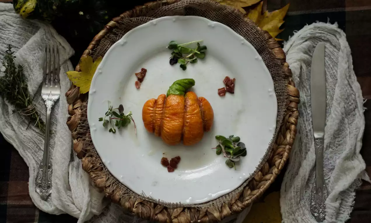 Dýňová buchta v tvare tekvice s oranžovou textúrou na bielom tanieri, ozdobená listom bazalky, mikrobylinkami a kúskami slaniny