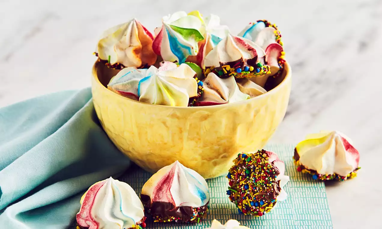 Colourful meringue kisses with swirls of pink, blue, green, and yellow, dipped in chocolate and sprinkles, arranged in a yellow bowl