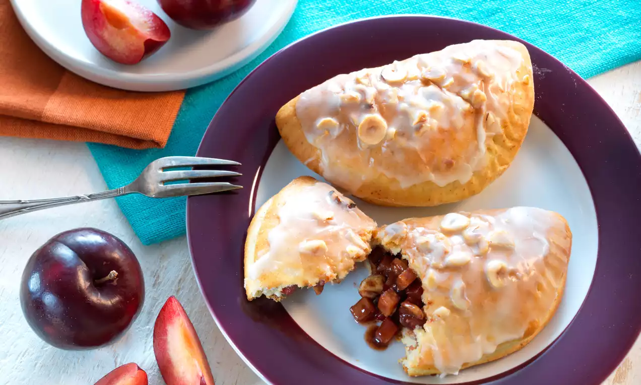Zwetschgentaschen auf Holzbrett, goldbrauner Blätterteig, glänzende dunkellila Pflaumenfüllung, gehackte Haselnüsse und Puderzucker