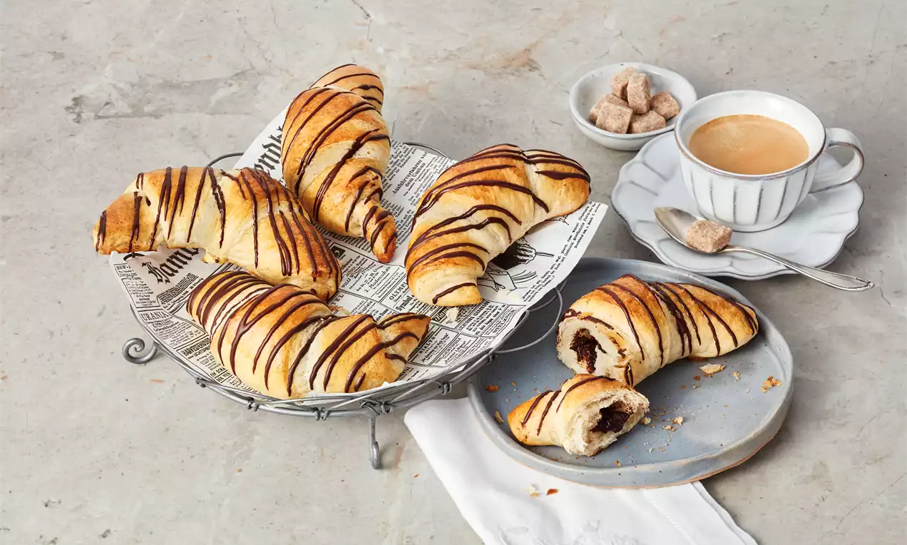 Goldbraune Schoko-Croissants mit dunklen Schokoladenstreifen auf Zeitungspapier in einem Metallkorb, daneben Kaffee und Zucker auf hellem Geschirr