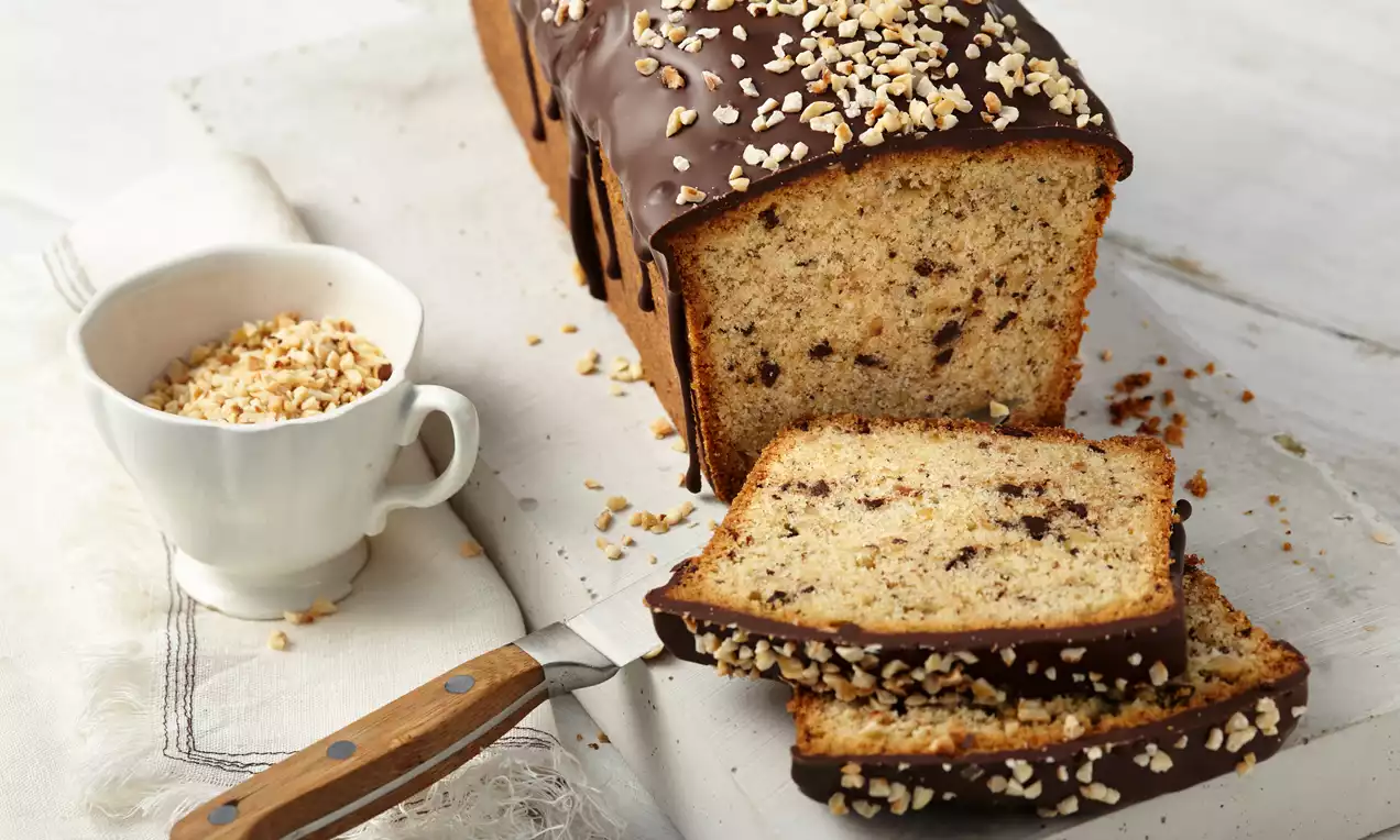 Saftiger Rührkuchen mit goldbrauner Krume, dunklen Schokostückchen und Nusskruste, teils aufgeschnitten, auf weißer Platte mit Messer