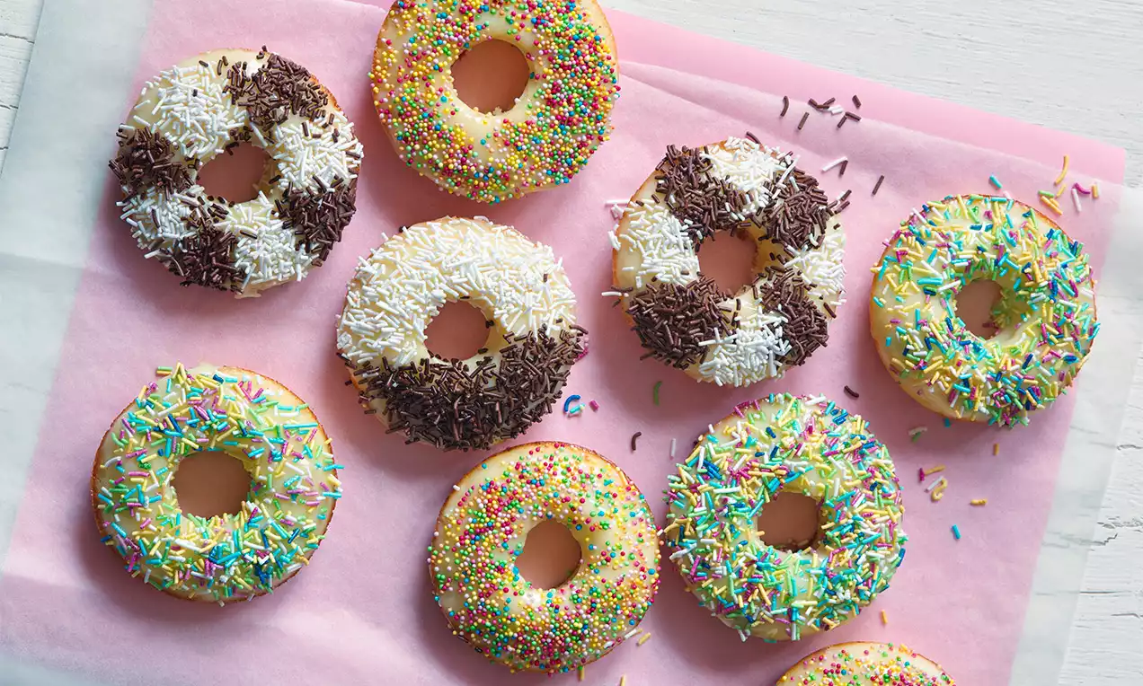 Pečené donuty zlatisté s jemne nadýchaným povrchom zdobené pestrými dúhovými a čokoládovými posýpkami, uložené na ružovom papieri