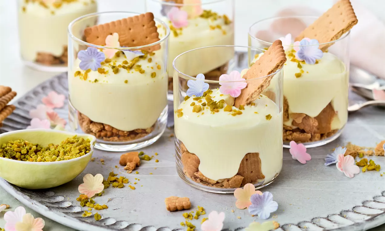 Zitronendessert in Gläsern mit gelber Creme auf Keksboden, garniert mit Pistazien, Butterkeksen und pastellfarbenen Zuckerdekoren
