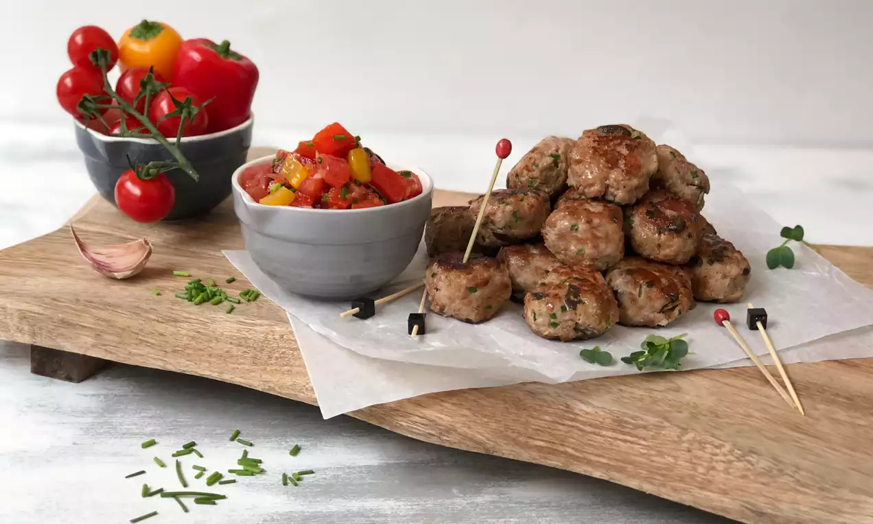 Ein Stapel goldbrauner Mini-Frikadellen auf weißem Pergamentpapier, daneben bunte Tomaten-Paprika-Salsa in einer grauen Schale auf Holzbrett