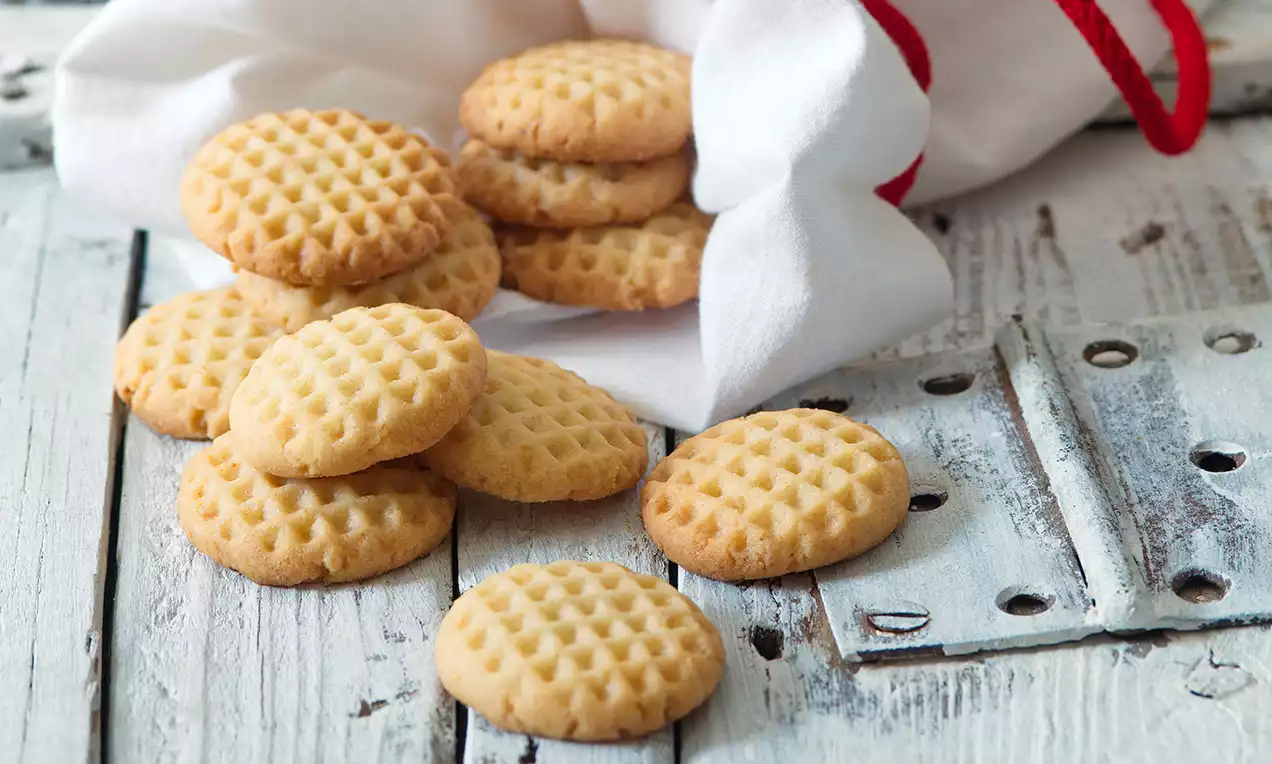 Citronové máslové sušenky světle zlaté barvy s jemným vzorem mřížky, volně rozložené na bílém dřevěném stole vedle látkového sáčku