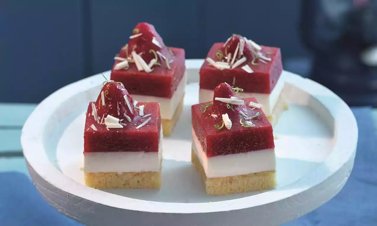 Četvrtasti kolači s bež biskvitom, bijelim slojem kreme i crvenim prelivom, ukrašeni jagodama, bijelom čokoladom i limetinom koricom