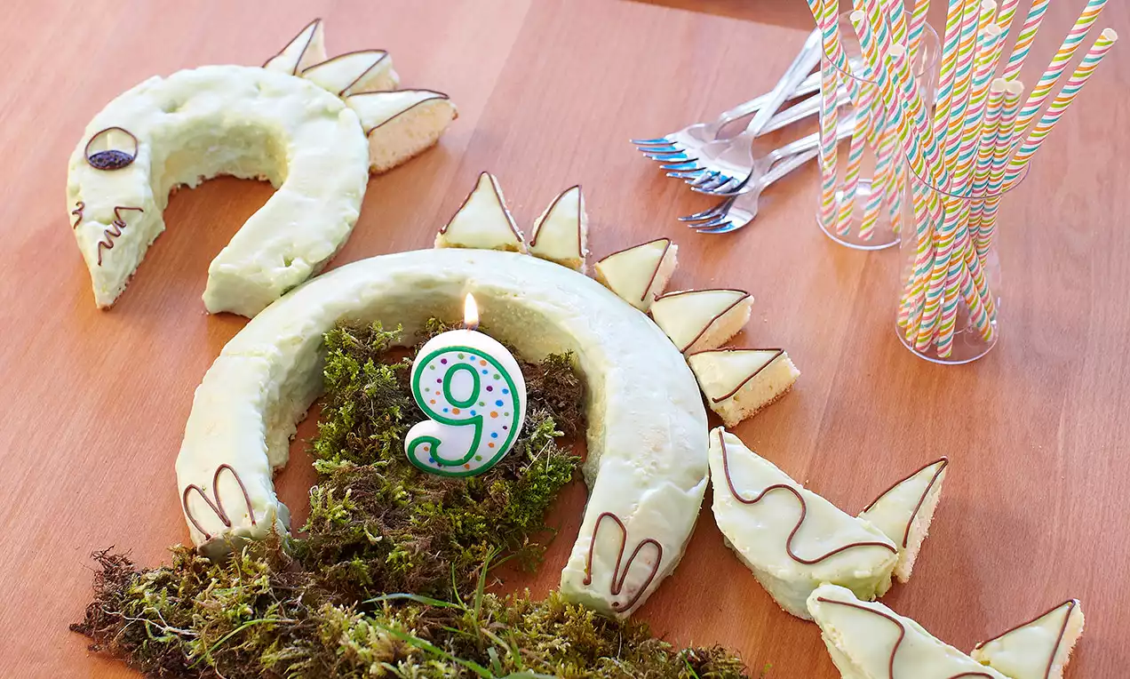 Torta verde chiaro a forma di dinosauro con decorazioni di cioccolato scuro, dettagli triangolari, muschio verde e candela numero 9 al centro