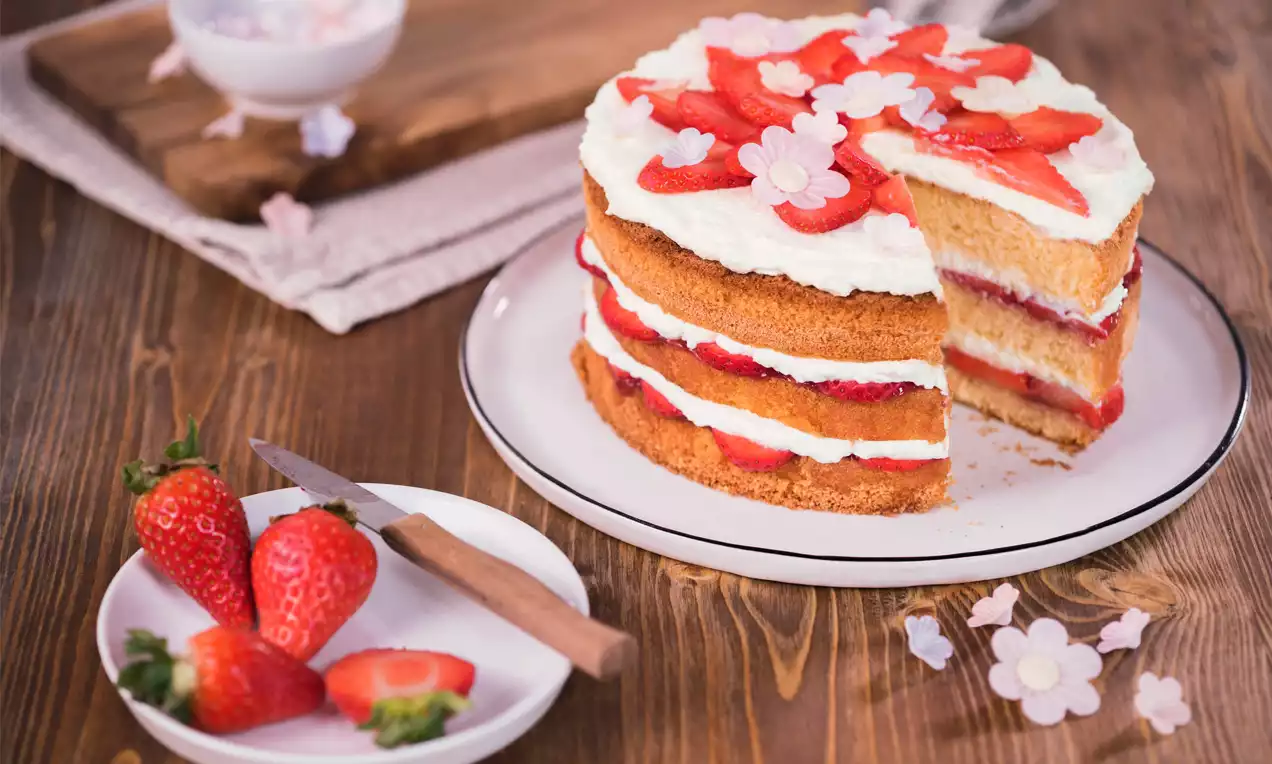 Dreischichtiger Sponge Cake mit hellbraunem Biskuit, weißer Creme, frischen roten Erdbeeren und Zuckerdekor auf einem weißen Teller