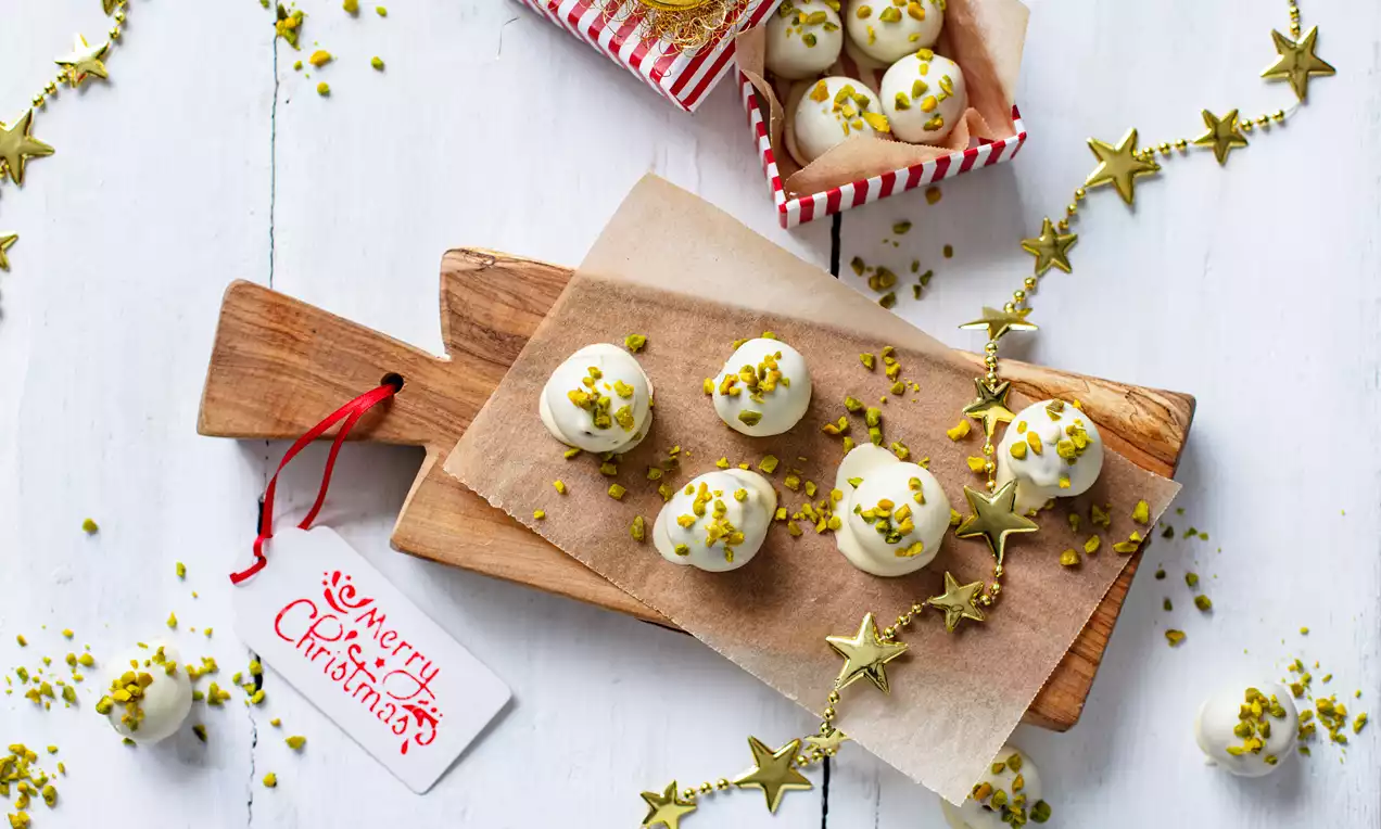 Lumenvalkoiset, pyöreät tryffelit siististi aseteltuina voipaperille puiselle tarjoilualustalle, koristeltuina kulta-vihreillä pistaasimuruilla