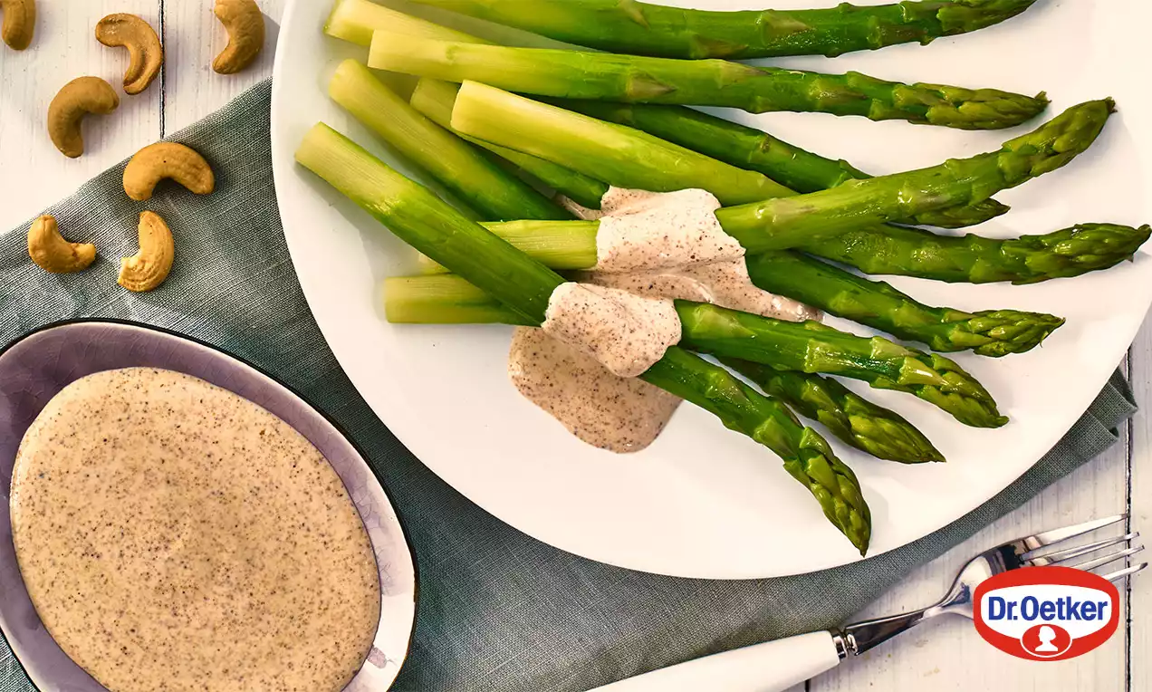 Štíhlé zelené chřestové výhonky na bílém talíři s jemnou světle hnědou kešu omáčkou, vedle misky s omáčkou a pár kešu oříšků