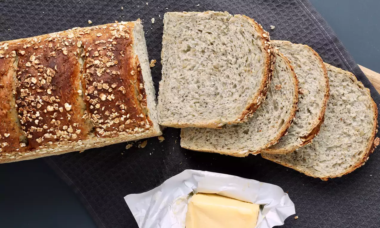 Vaaleanruskean kuoren ja kaurahiutaleiden peittämä vuokaleipä viipaleina tummaa kangasta vasten, vieressä avattu voi kääreessä