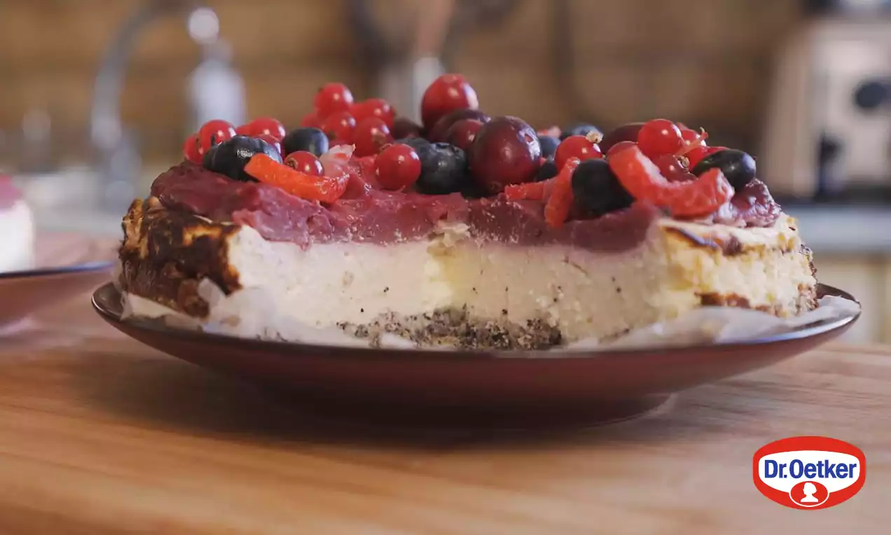 Makový cheesecake s vrstvenou štruktúrou svetlej plnky a tmavého maku, na vrchu lesklá červená ovocná poleva s bobuľami na hnedastom tanieri