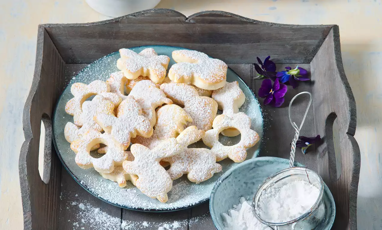 Tvarohové sušenky světle zlaté barvy s jemným moučkovým cukrem, ve tvarech květin a zajíčků, naaranžované na modrém talíři s fialovými kvítky