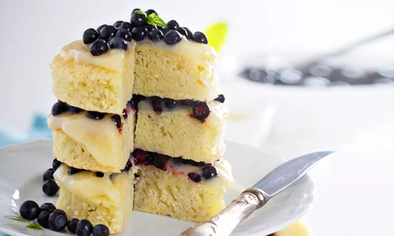 Stacked moist vanilla cake slices with pale yellow glaze, scattered blueberries on top and between layers, served on a white plate