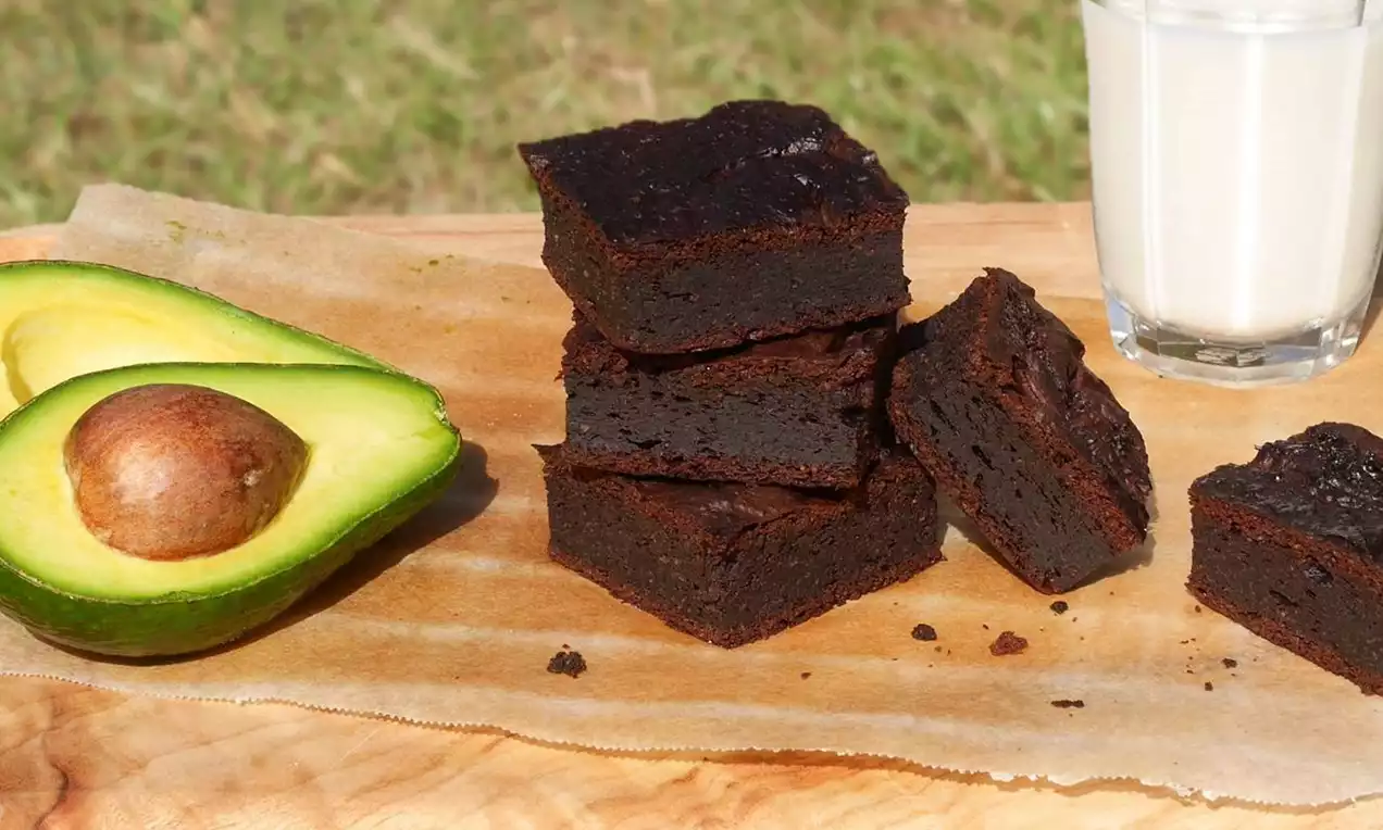 Brownies densi e scuri con una superficie leggermente lucida, impilati su carta forno vicino a metà avocado verde brillante e un bicchiere di latte