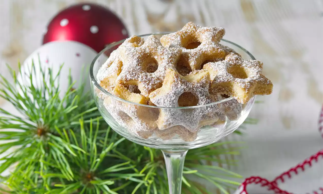 Zlatisté hviezdičkové sušienky s jemným práškovým cukrom, uložené v čírom sklenenom pohári, vedľa vetvičky ihličnanu a vianočných gulí
