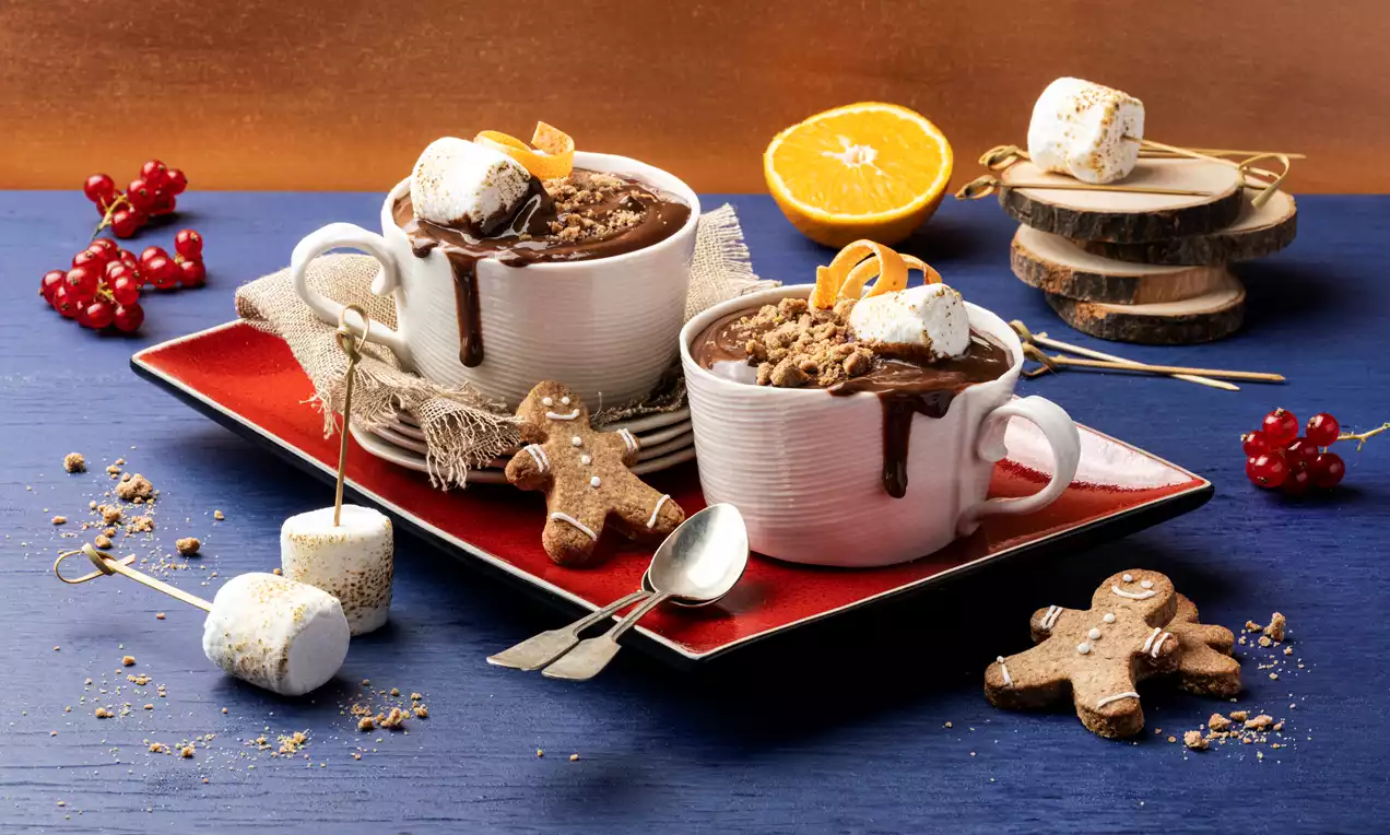 Tazze bianche colme di cioccolata cremosa decorata con marshmallow dorati, granella croccante e spicchi d’arancia su un vassoio rosso