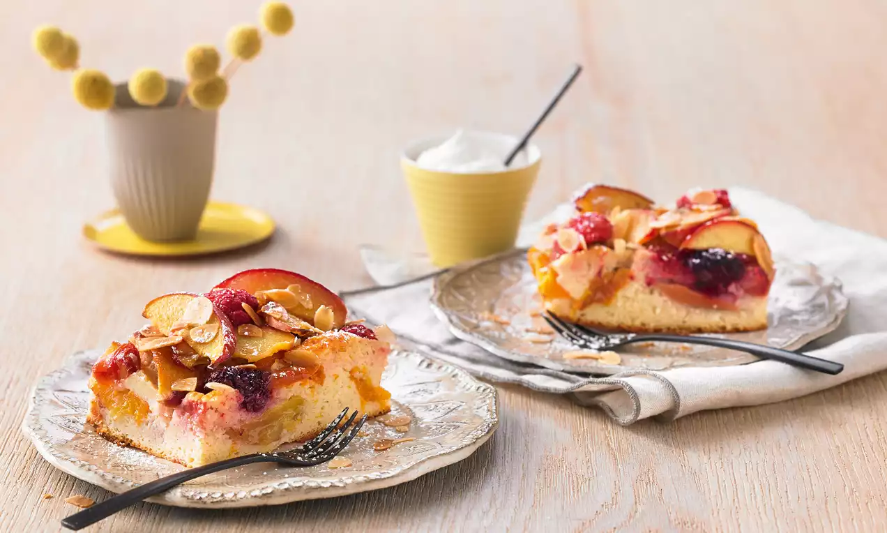 Zwei Stück sommerlicher Obstkuchen mit goldgelbem Rührteig, fruchtigem Belag und Mandelblättchen auf geschwungenen Glastellern angerichtet