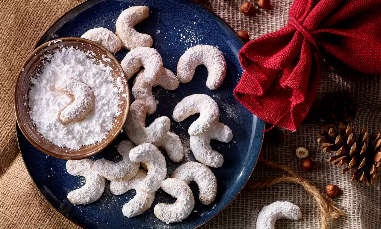 Vanillekipferl in gebogener Form, goldbraun unter dichtem Puderzucker, arrangiert auf einem blauen Teller neben Schale mit Puderzucker