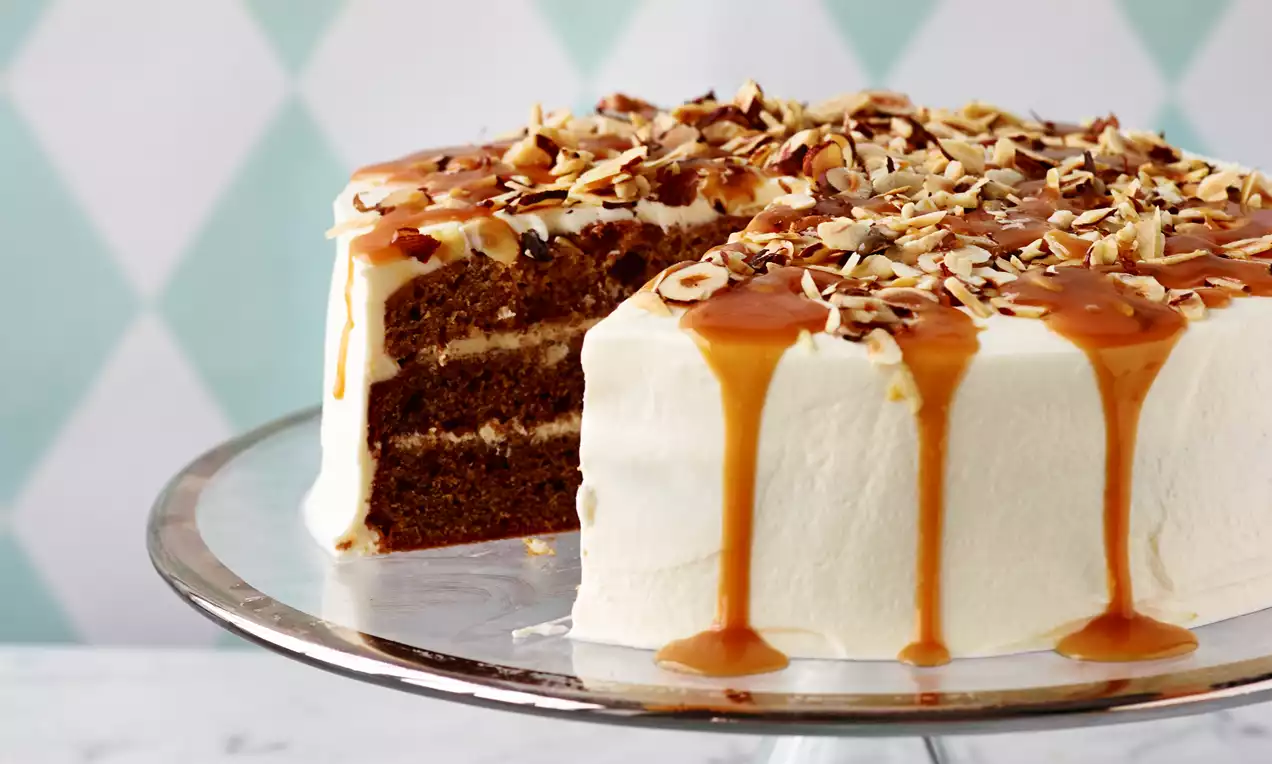 Høj lagkage med hvid flødeskumsoverflade, karamelsauce der løber ned, og drys af hakkede nødder serveret på et sølvfad