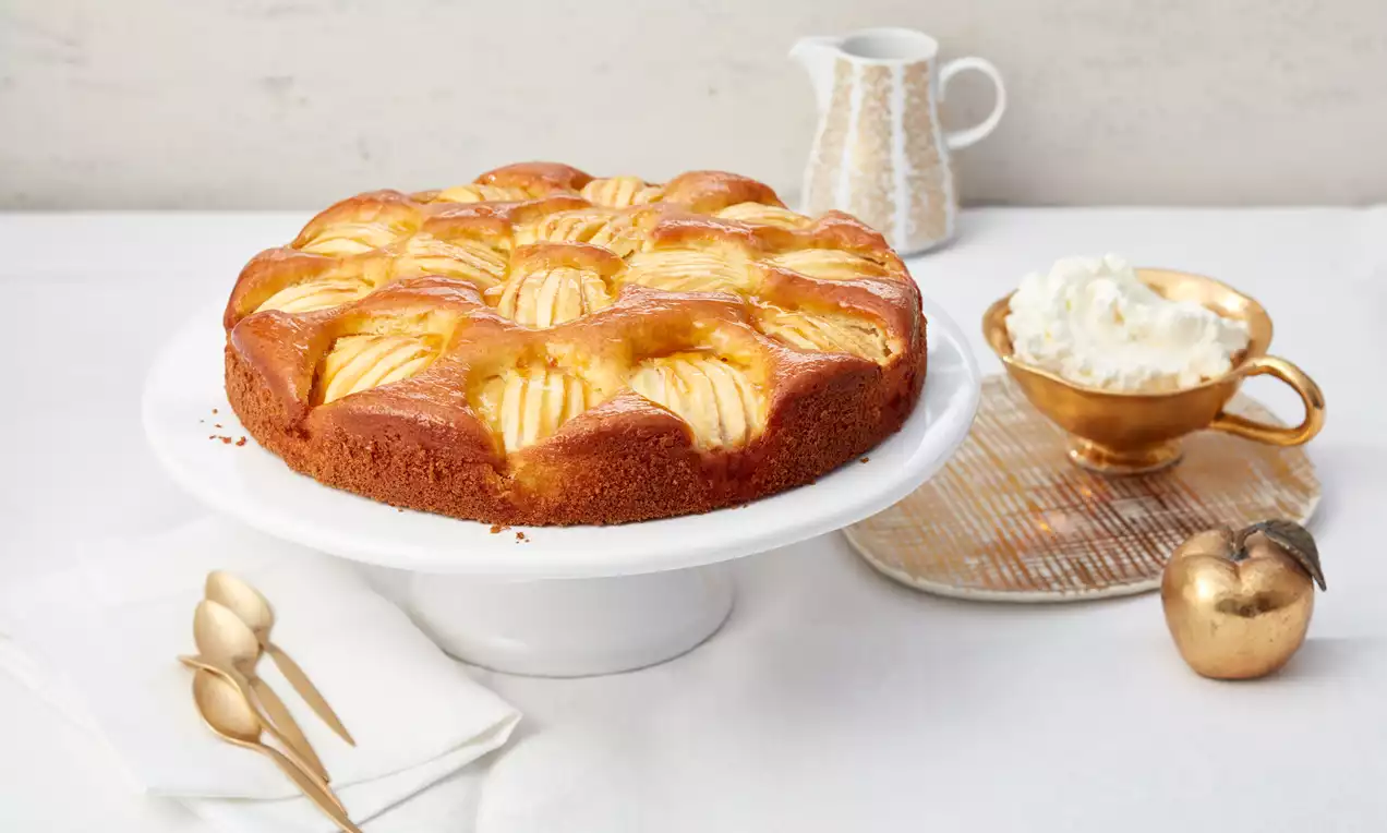 Karamelize elma dilimlerinin altın tonlarında parladığı kabarık ve nemli elmalı kek, beyaz bir servis tabağında sunulmuş